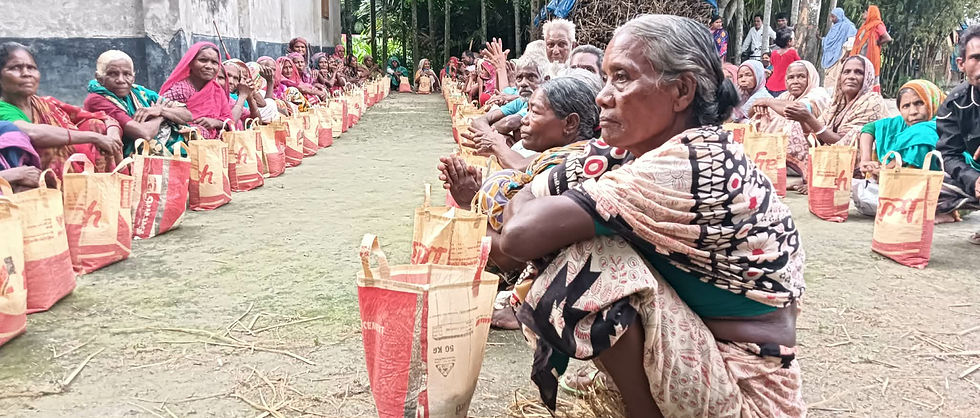 A large group of rural villagers, mostly elderly women, sit in two lines outside a simple building, each with a bag of food supplies. A woman in a patterned sari sits in the foreground, while trees and a thatched shelter surround the gathering.