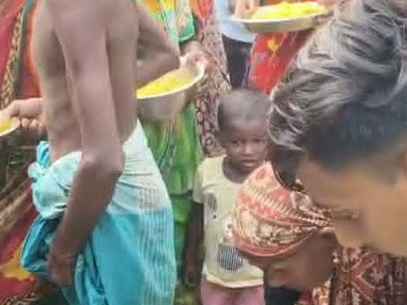 A young volunteer bends down to serve food to an elderly community member, as a crowd of people wait patiently with bowls of yellow rice, and a small child watches quietly in the background.