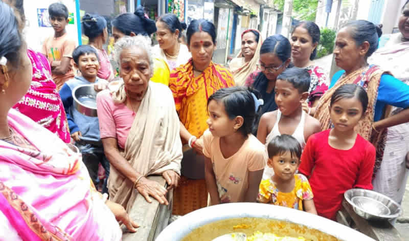 A group of people, primarily women and children, gathered together in what appears to be a community setting. An elderly woman in light-colored clothing sits in the center, surrounded by women wearing colorful traditional garments in pink, orange, and other bright colors. Several children of various ages are present, including young girls in the foreground. The scene suggests a community gathering or social event, with people appearing to be engaged in conversation or participating in some type of community activity.