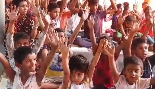 A large group of children sitting together with their hands raised enthusiastically in the air. The children appear to be of various ages, wearing casual clothing in different colors including white, orange, and other bright hues. They seem engaged and excited, possibly participating in a classroom activity, community event, or educational program. The setting appears to be indoors with natural lighting