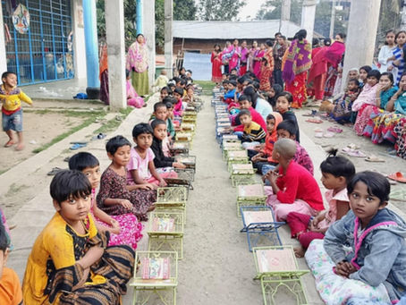 A large group of children and adults gathered in an outdoor courtyard for what appears to be a community meal or food distribution event. Dozens of children, ranging from toddlers to teenagers, are seated in organized rows on the ground with green metal serving trays or plates placed in front of them. The children are wearing colorful traditional Indian clothing including saris, kurtas, and other garments in bright yellows, pinks, reds, and other vibrant colors.