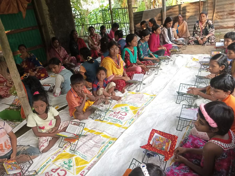 A large group of children and women sitting cross-legged on white sheets laid on the ground beneath a shaded outdoor area, attending an open-air community class or gathering. Small wire book stands holding books and papers are arranged in front of the children along the length of the sheet. Green vegetation and a fence are visible in the background, suggesting a rural setting.
