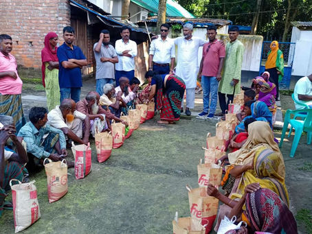 A community distribution event taking place in an outdoor courtyard area with brick buildings in the background. A group of officials or organizers in formal attire (shirts, some in traditional white kurtas) stand together in the center-back of the scene. Seated on the ground in organized rows are community members, many wearing colorful traditional clothing and head coverings, including women in saris and head scarves in various bright colors like yellow, purple, red, and blue. Each person has a paper bag or package placed in front of them on the ground. Green plastic chairs are visible on the right side. The setting appears to be a rural or semi-urban community area with simple brick and concrete structures
