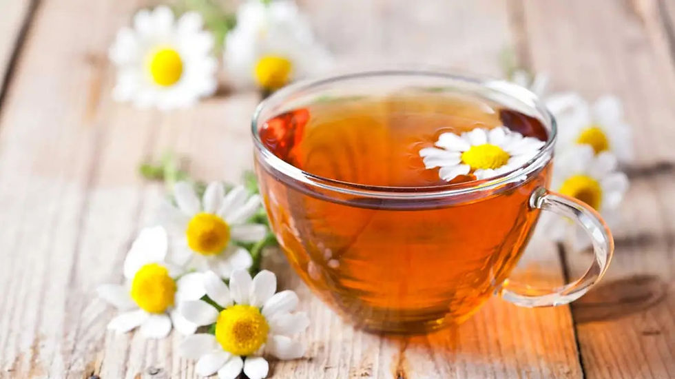 A clear glass teacup filled with golden amber chamomile tea sits on a rustic wooden surface. Fresh chamomile flowers with white petals and bright yellow centers are scattered around the cup, with one flower floating on the surface of the tea.