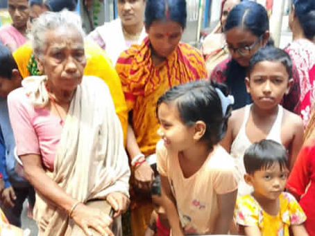 A group of people, primarily women and children, gathered together in what appears to be a community setting. An elderly woman in light-colored clothing sits in the center, surrounded by women wearing colorful traditional garments in pink, orange, and other bright colors. Several children of various ages are present, including young girls in the foreground. The scene suggests a community gathering or social event, with people appearing to be engaged in conversation or participating in some type of community activity.