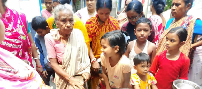 A group of people, primarily women and children, gathered together in what appears to be a community setting. An elderly woman in light-colored clothing sits in the center, surrounded by women wearing colorful traditional garments in pink, orange, and other bright colors. Several children of various ages are present, including young girls in the foreground. The scene suggests a community gathering or social event, with people appearing to be engaged in conversation or participating in some type of community activity.