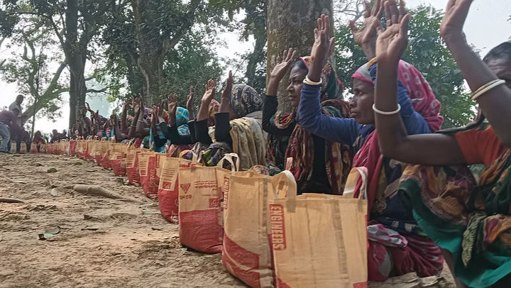 A long row of community members, mostly women, sit outdoors along a dirt path beneath large trees, each with a filled burlap bag of rice in front of them. Many raise their hands, appearing to respond to a question or express gratitude during a food aid distribution event. 