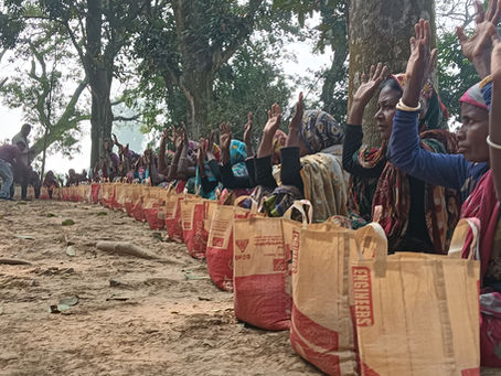 A long row of women sit on the ground outdoors under trees, each with a filled burlap/woven bag of rice in front of them. Many raise their hands, appearing to express gratitude or acknowledgment during a community rice distribution event. You said: please write a caption for those images