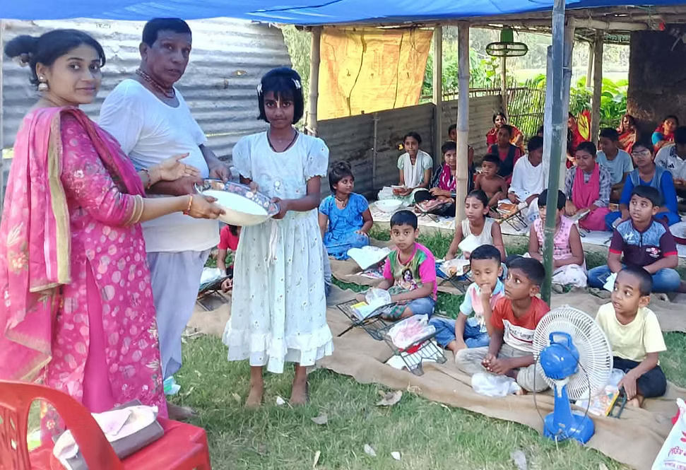 Two adults handing a bowl of food to a young girl standing in an outdoor classroom setup.