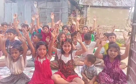 A group of approximately 30-40 children and young people sitting on the ground in what appears to be a courtyard or outdoor area between buildings. Most of the children have their hands raised up in the air, appearing happy and enthusiastic. They are wearing a mix of colorful clothing including traditional-style dresses, casual shirts, and various bright colors like red, purple, green, and white. The setting looks like it could be in a developing community, with simple concrete block buildings visible in the background. The children appear to be participating in some kind of group activity or responding to someone off-camera, creating a joyful, communal atmosphere