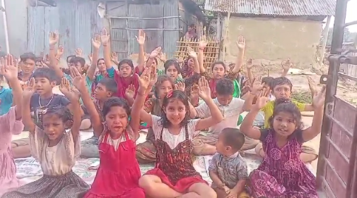 A group of approximately 30-40 children and young people sitting on the ground in what appears to be a courtyard or outdoor area between buildings. Most of the children have their hands raised up in the air, appearing happy and enthusiastic. They are wearing a mix of colorful clothing including traditional-style dresses, casual shirts, and various bright colors like red, purple, green, and white. The setting looks like it could be in a developing community, with simple concrete block buildings visible in the background. The children appear to be participating in some kind of group activity or responding to someone off-camera, creating a joyful, communal atmosphere.