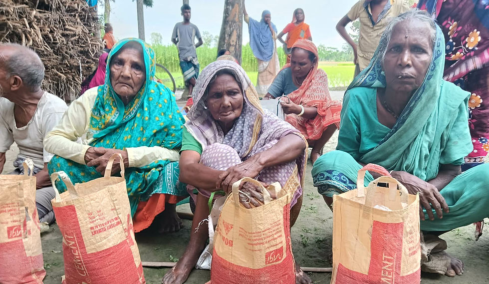 "Three elderly women sit on the ground in a rural Bangladesh village, each with a large filled food relief bag in front of them, with an elderly man and other community members visible behind them beside lush green rice fields."