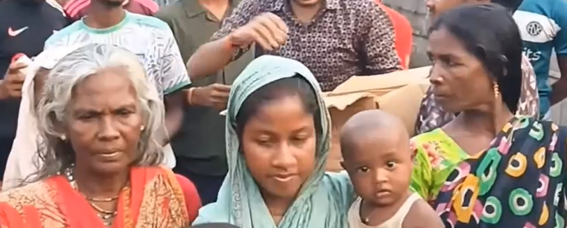 "An elderly woman, a young mother holding a toddler, and another woman stand together in a crowd during a community relief event in Bangladesh, with volunteers and other recipients visible in the background."