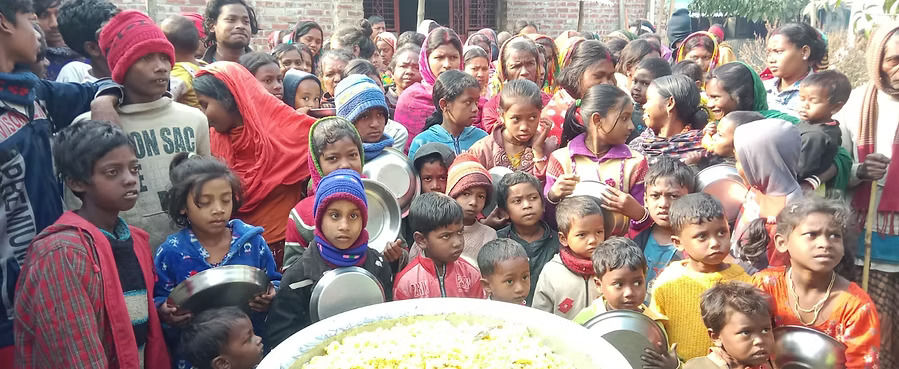 A large crowd of children and women in warm winter clothing gather with metal plates and bowls around a massive pot of khichdi during an outdoor community food distribution event.