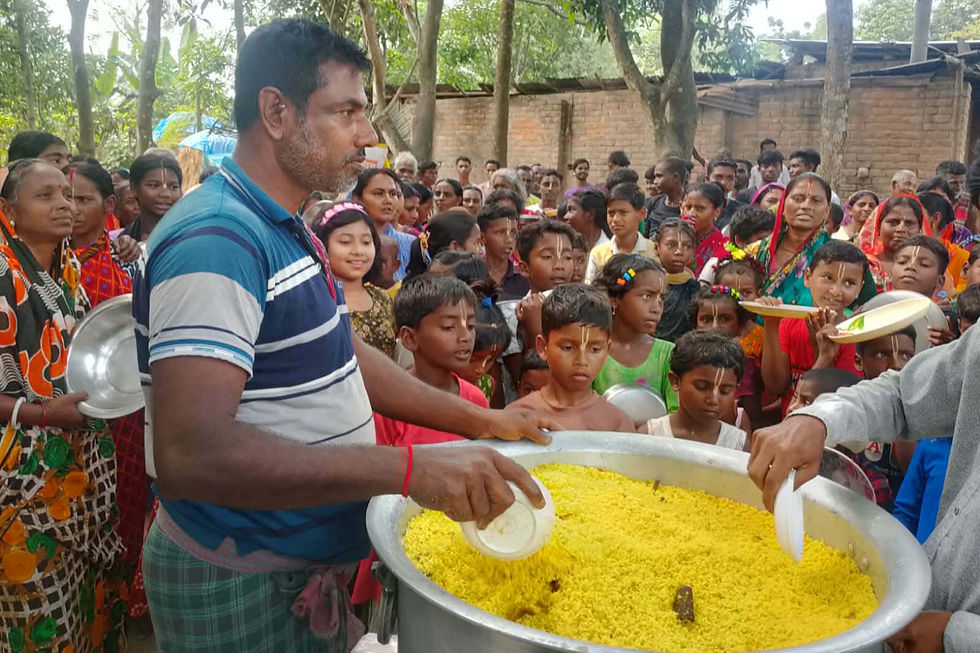 A man in a blue striped polo shirt serves bright yellow khichri (a rice and lentil dish) from a large aluminum pot to a crowd of villagers, including children and women in colorful saris, who hold out steel plates to receive food at an outdoor community meal distribution event, with trees and a brick wall in the background.