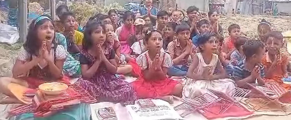 A group of young children sitting cross-legged outdoors, chanting with palms folded together in a prayer gesture in front of small study stands.