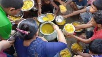 A group of people gathered in a circle on the ground, sharing a communal meal from metal plates and bowls containing yellow rice or dal. Hands reach toward the center as multiple individuals participate in this community dining experience.