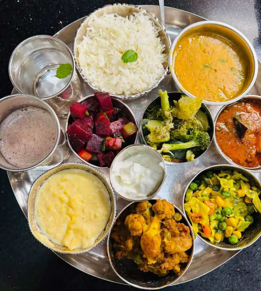 An elaborate Indian thali served on a large stainless steel plate with multiple small bowls arranged in a circular pattern. The meal includes white basmati rice garnished with mint leaves in the center, surrounded by various dishes: a golden yellow dal or curry soup, orange-red curry (possibly butter chicken or paneer makhani), diced red beetroot salad, mixed green vegetables with broccoli and other greens, yellow mixed vegetables or sabzi, white yogurt or raita, creamy yellow curry or kheer, and what appears to be cauliflower or potato curry. There's also a glass of water and what looks like a chocolate-colored drink or soup. The diverse array of colorful dishes represents a complete traditional Indian meal with vegetables, legumes, dairy, and various spiced preparations arranged in the classic thali style.