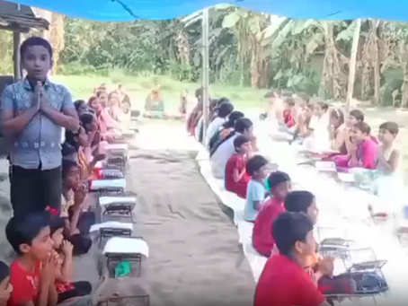 Children sit in rows on the ground under a blue tarp canopy in an outdoor setting, each with a metal plate in front of them, waiting to be served a meal. A woman stands at the front left holding a microphone, appearing to lead the gathering. The scene takes place in a rural area with banana trees and simple structures visible in the background.