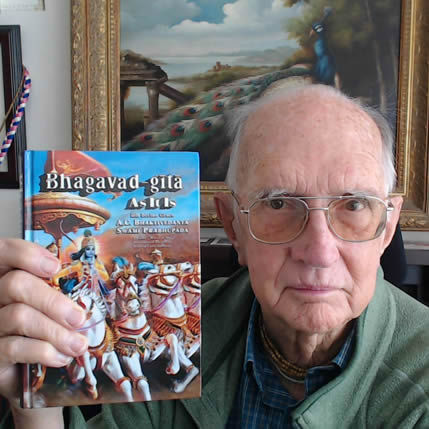 An elderly man with glasses wearing a green sweater over a blue plaid shirt holds up a copy of the Bhagavad-gita As It Is book, which features colorful cover art depicting Krishna and Arjuna on a chariot. Behind him is a framed painting of a peacock in a pastoral landscape with classical architecture.