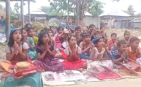 Children sitting on colorful mats and blankets outdoors in what appears to be a makeshift classroom or learning space. The group consists of approximately 20-25 children of various ages, sitting cross-legged on red, green, and other colored fabric. They appear to be in a rural or village setting with simple buildings and trees visible in the background. Some children are holding books or papers, suggesting this may be an outdoor educational activity or community learning session. The children are dressed in casual clothing and appear engaged and attentive.