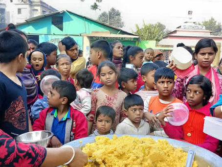 A community food distribution event where a large group of children and adults have gathered around a table with a large tray of kitchri. A person in dark clothing with red patterns is serving the food using a ladle from a metal container. The crowd includes many children of various ages wearing colorful clothing - red, blue, pink, and other bright colors. Some people are holding plates or containers to receive food. The setting appears to be outdoors in a rural area with houses and a green-roofed structure visible in the background. The scene depicts a community meal service or food assistance program.