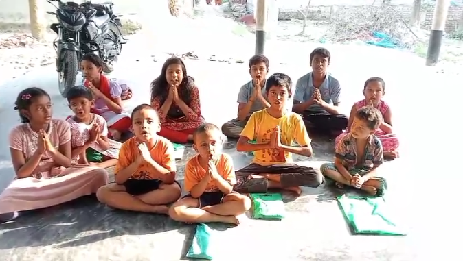 A group of children of various ages sitting cross-legged on the ground in what appears to be an outdoor learning environment. The children are wearing colorful clothing including orange, pink, and other bright colors. They have notebooks and educational materials spread in front of them on the ground. In the background, there are motorcycles parked and what looks like a simple building structure. The setting appears to be informal, possibly a community-based education program or outdoor classroom taking place in a rural or semi-urban area. The children appear attentive and engaged in their learning activity.