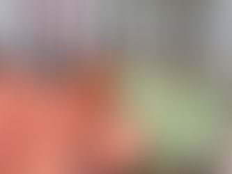 A group of women and children stand in a line at an outdoor public monument during dusk, holding blankets in shades of pink, peach, and light green. The individuals are wearing traditional South Asian clothing including colorful saris and head coverings. Behind them is a white modern monument structure with geometric frames and a tower-like element. The setting includes palm trees, residential buildings, and a paved plaza area with red and white striped steps. Other people can be seen in the background. The scene appears to be documenting a blanket distribution event at a community gathering space.