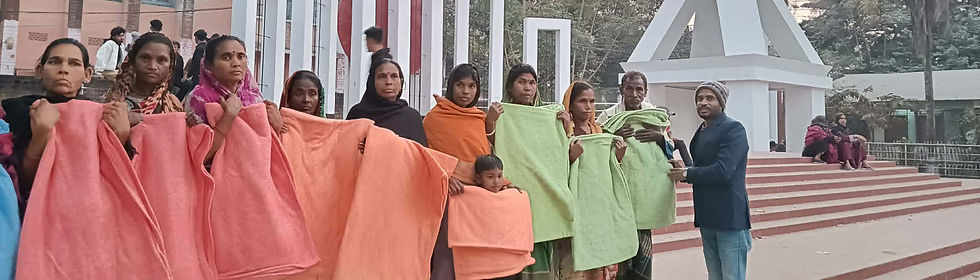 A group of women and children stand in a line at an outdoor public monument during dusk, holding blankets in shades of pink, peach, and light green. The individuals are wearing traditional South Asian clothing including colorful saris and head coverings. Behind them is a white modern monument structure with geometric frames and a tower-like element. The setting includes palm trees, residential buildings, and a paved plaza area with red and white striped steps. Other people can be seen in the background. The scene appears to be documenting a blanket distribution event at a community gathering space.