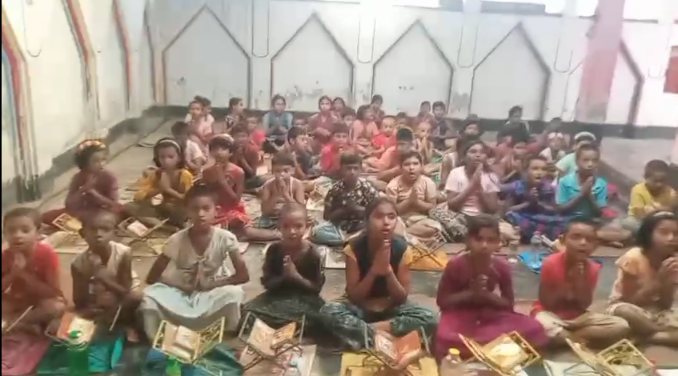 A large group of children and young people sitting cross-legged on the floor in what appears to be a classroom or community center. The participants are wearing colorful clothing in various shades including red, yellow, blue, and green. Many have books or educational materials in front of them. The room has white walls with decorative house-shaped designs or cutouts visible in the background. The setting suggests this is likely an educational program, literacy class, or community learning session taking place indoors.RetryClaude can make mistakes. Please double-check responses. Sonnet 4
