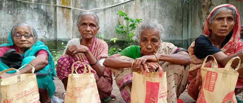 Four elderly women sitting together outdoors against a weathered concrete wall with some greenery visible in the background. Each woman is holding a light-colored paper or cloth shopping bag. From left to right: a woman wearing glasses and a teal/turquoise sari, a woman in a pink/red patterned sari, a woman in a light-colored sari, and a woman wearing a red and white head covering. The setting appears to be in a courtyard or outdoor community space, suggesting this may be related to a distribution event or community program.