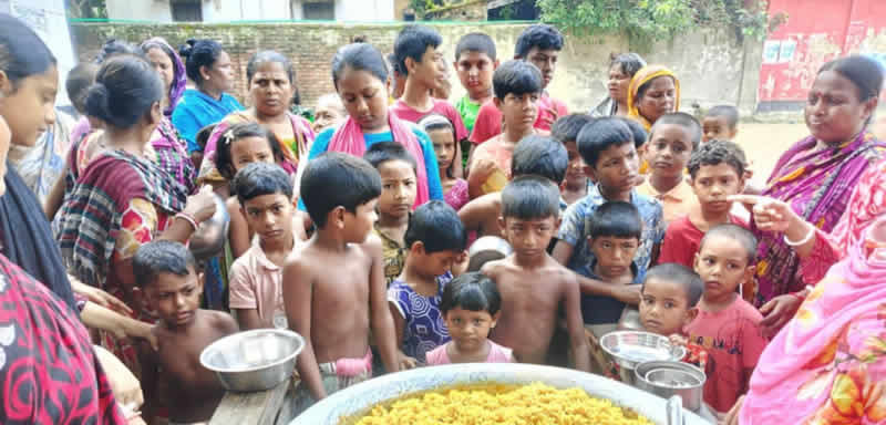 A large group of women and children gathered around a large metal pot or vessel containing Kichri. Many of the children are shirtless young boys, and there are women wearing colorful traditional clothing including saris in various colors like pink, green, and patterned fabrics. Some people are holding metal plates or bowls. The setting appears to be outdoors in a community area with brick buildings visible in the background. This looks like a community meal distribution or feeding program, with the crowd focused on the large serving vessel in the center.