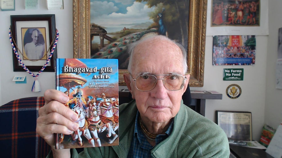 An elderly man with glasses and thinning gray hair sits in what appears to be a home office or study, wearing a green cardigan over a plaid shirt. He is holding up a copy of the Bhagavad Gita ("Bhagavad-gita As It Is" by A.C. Bhaktivedanta Swami Prabhupada) which features colorful artwork depicting Krishna and Arjuna on a battlefield with horses and chariots. Behind him on the walls are various framed pictures including what appears to be a portrait of a spiritual figure, landscape paintings, and small decorative items. There's also a "No Farms No Food" sign visible among the wall decorations. The setting suggests someone with interests in spirituality, philosophy, and possibly agriculture or environmental causes.