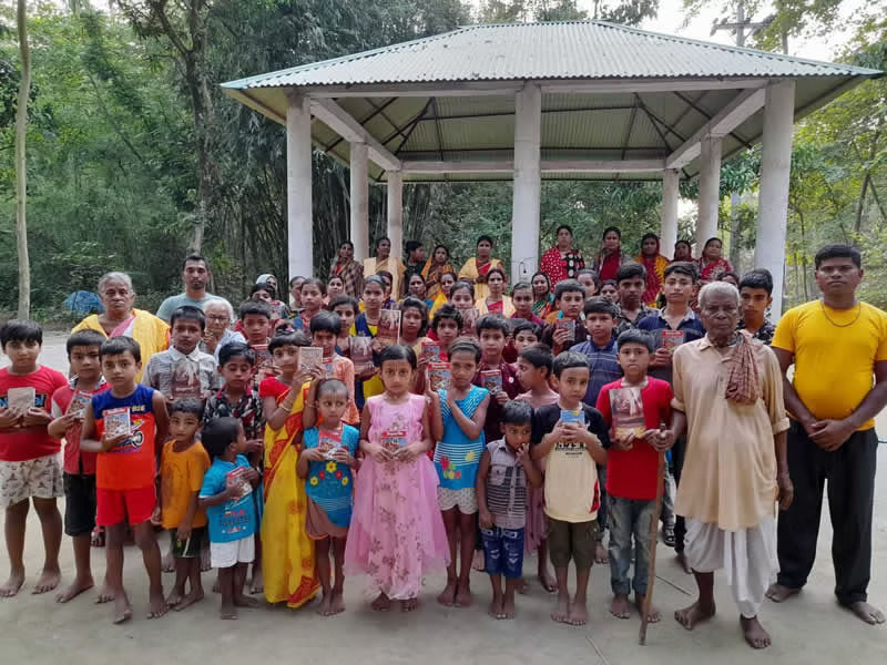 "A large group of children and adults stand together in front of an open-sided pavilion, many of the children holding books."