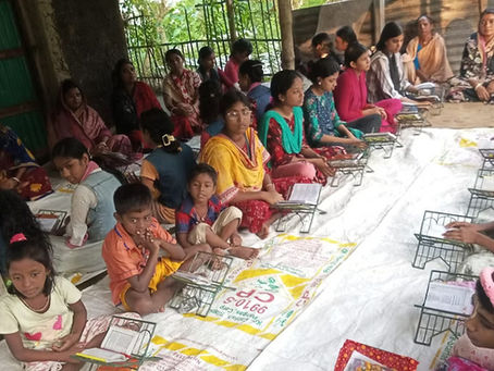 A group of children and adults seated on the floor in an open-air outdoor classroom, engaged in a Bhagavad Gita study session. Students are arranged in rows with small book stands holding scripture books in front of them. The setting appears to be a simple covered space with a concrete floor and lush greenery in the background.