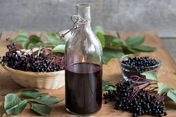 Clear glass swing-top bottle filled with dark elderberry syrup on a wooden surface. Fresh elderberries on their stems are displayed in a woven basket and glass bowl, with green elderberry leaves scattered around against a gray background.