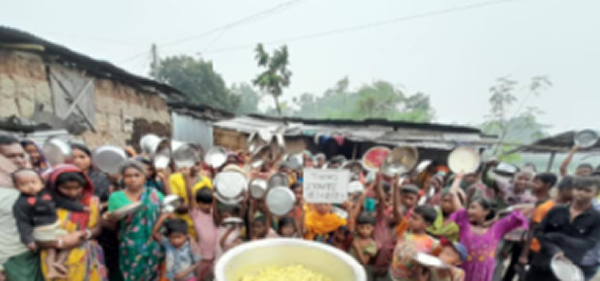 A large crowd of people, including many children and adults, gathered outdoors in what appears to be a community or village setting. The group is holding white plates or bowls, suggesting a community meal or food distribution event. In the foreground, there's a large yellow container or pot. The scene takes place in front of simple buildings with metal roofing, and trees are visible in the background under an overcast sky. The gathering appears to be a community food service or charitable meal distribution.