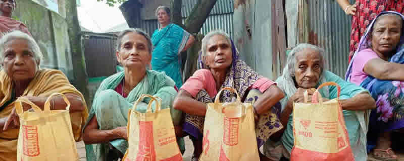A group of elderly women wearing traditional saris and head coverings sit together, each holding small paper bags or packages that appear to contain food or aid supplies. The women are seated in what looks like an outdoor distribution area, with more people visible in the background. This appears to be a food distribution or humanitarian aid event, likely in South Asia, where community members are receiving essential supplies.