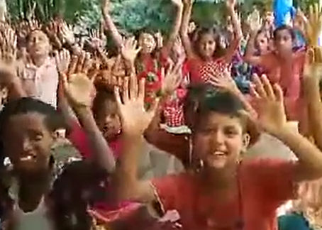 A large group of children sitting outdoors with their hands raised enthusiastically, smiling and cheering during what appears to be an outdoor classroom or community gathering, with green trees in the background.