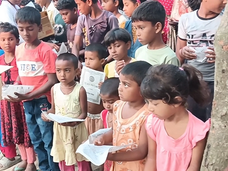 A long queue of young children standing outdoors near a tree, each holding a small printed paper or certificate, waiting to receive food items being distributed from a cardboard box by a volunteer's hand visible on the left. The children are dressed in casual colorful clothes, depicting a charitable treat or snack distribution event in a rural South Asian village setting.