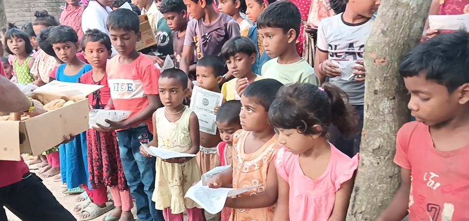 A long queue of young children standing outdoors near a tree, each holding a small printed paper or certificate, waiting to receive food items being distributed from a cardboard box by a volunteer's hand visible on the left. The children are dressed in casual colorful clothes, depicting a charitable treat or snack distribution event in a rural South Asian village setting.