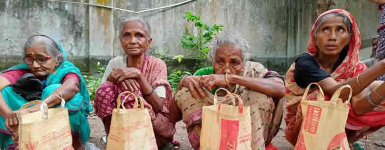 Four elderly women sitting together outdoors against a weathered concrete wall with some greenery visible in the background. Each woman is holding a light-colored paper or cloth shopping bag. From left to right: a woman wearing glasses and a teal/turquoise sari, a woman in a pink/red patterned sari, a woman in a light-colored sari, and a woman wearing a red and white head covering. The setting appears to be in a courtyard or outdoor community space, suggesting this may be related to a distribution event or community program.