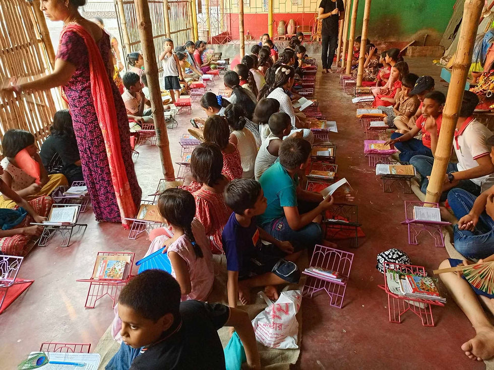 A large group of children sit on the floor of an open bamboo-structured classroom in Bangladesh, each with a small metal book stand and reading materials in front of them, as a female teacher in a red sari oversees the lesson.