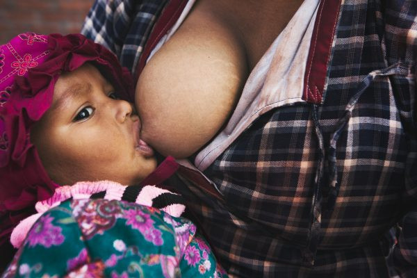 A Black mother breastfeeding. The mother is wearing a tartan feeding top in burgundy, black, and white. The baby wears a floral outfit in pinks and greens.