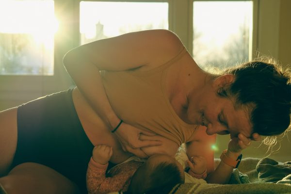 A white mother breastfeeding while lying down. The mother wears a beige vest and black underwear. The baby wears a neutral babygrow. The sun shines in through a window behind them.