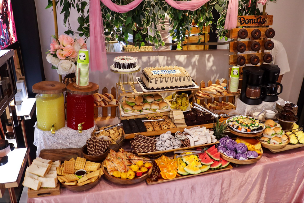 A photo of a food table holding various food selections such as cake, drink refreshments, cookies, and bite-size sandwiches.