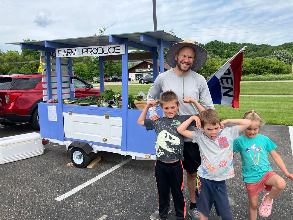 Farmer David and his three kids pose flexing by a blue farm produce stand at the New Glarus Farmers Market held in a parking lot under a bright sky.