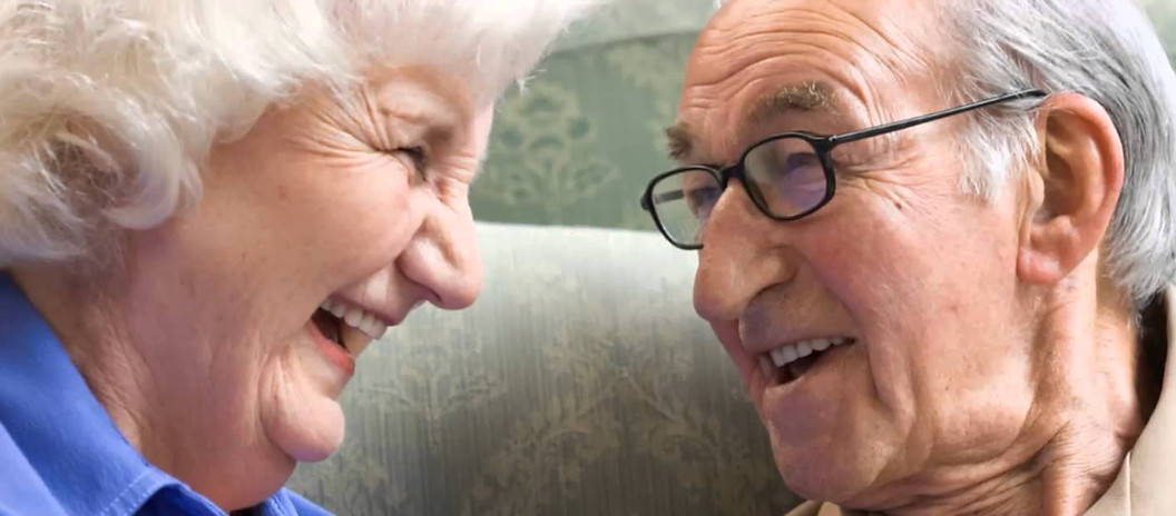 An elderly couple, relaxing on their sofa and laughing.
