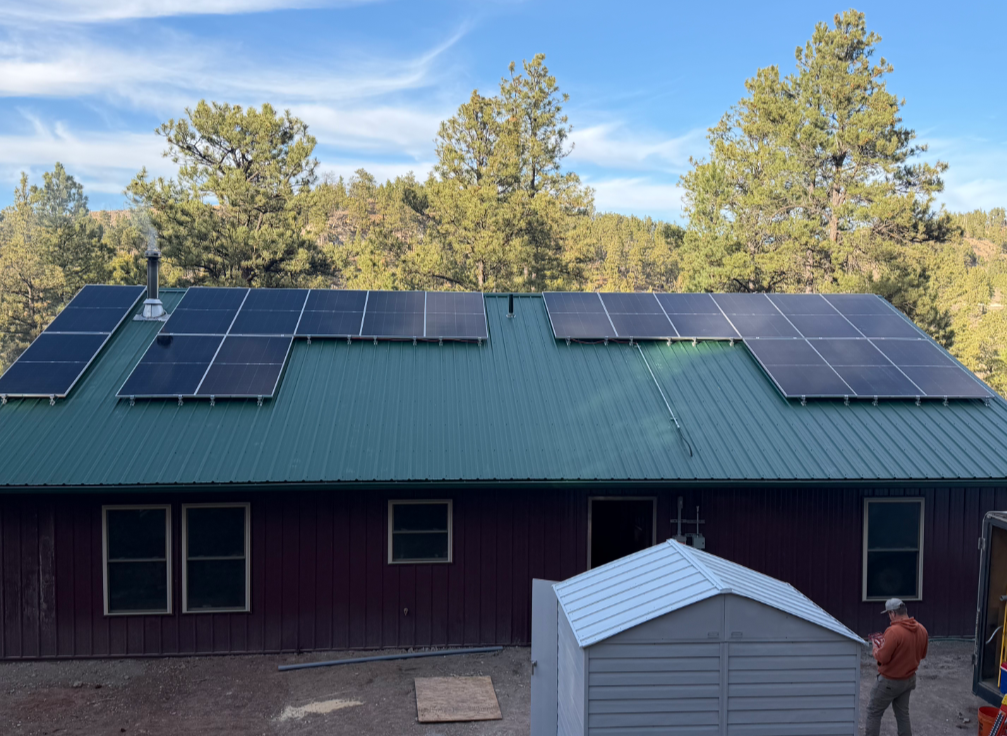 Solar panels installed across two sections of a green metal roof, with trees in the background
