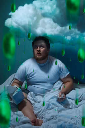 Conceptual lifestyle photography of sick man sitting under a cloud inside with nyquil pills raining down on him. Under the weather visual pun.